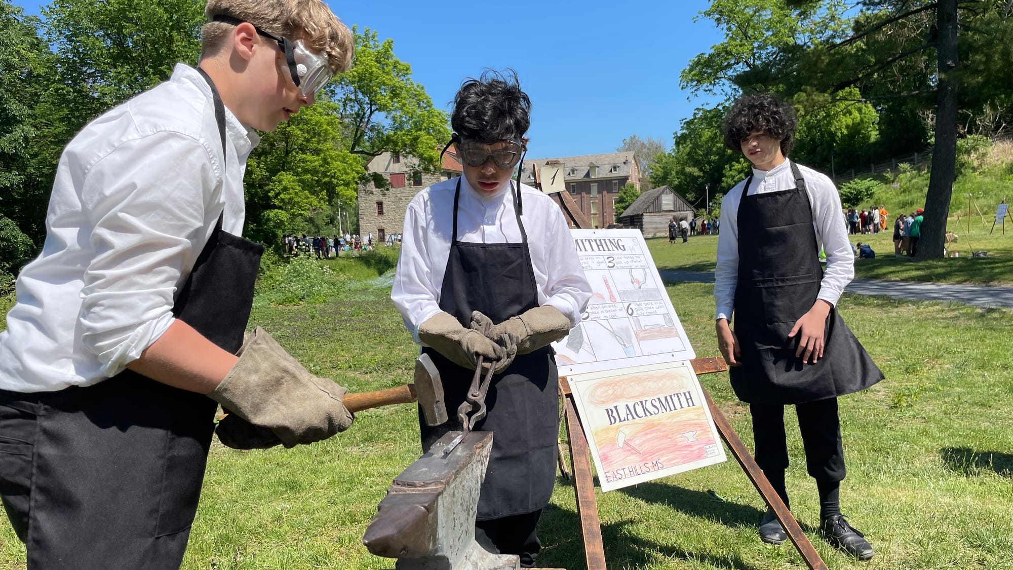 Aiden Monteiro hammers while Heinz Schmitt holds the hot piece of metal steady. Wyatt Cole speaks to students on the blacksmi