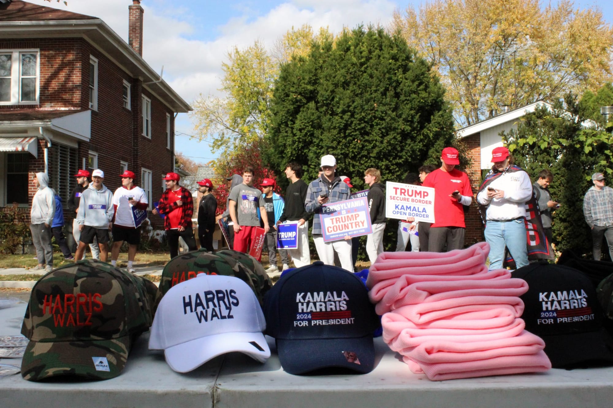 Donald Trump supporters stand in the background of a merchandise table selling hats and other items for the Harris-Walz campa